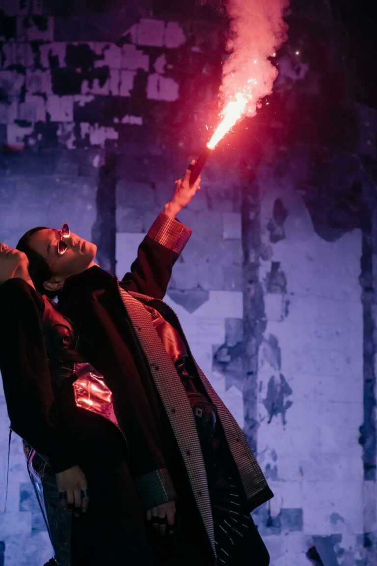Two individuals in stylish attire holding a flare in a moody urban environment.
