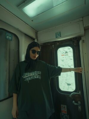 Woman in dark clothing on train, wearing sunglasses and hijab, with graffiti-inspired design on shirt.